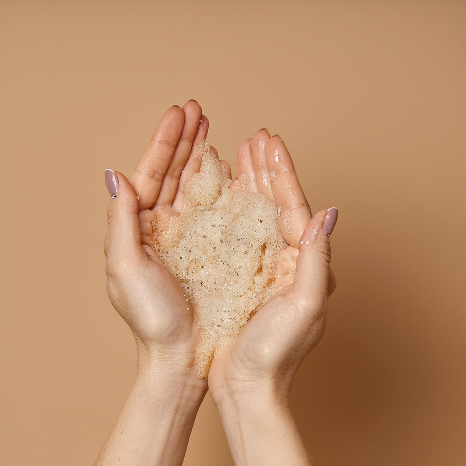 a view of women’s hands full of creamy scrub. Behind the hands there is an endless, solid beige background.