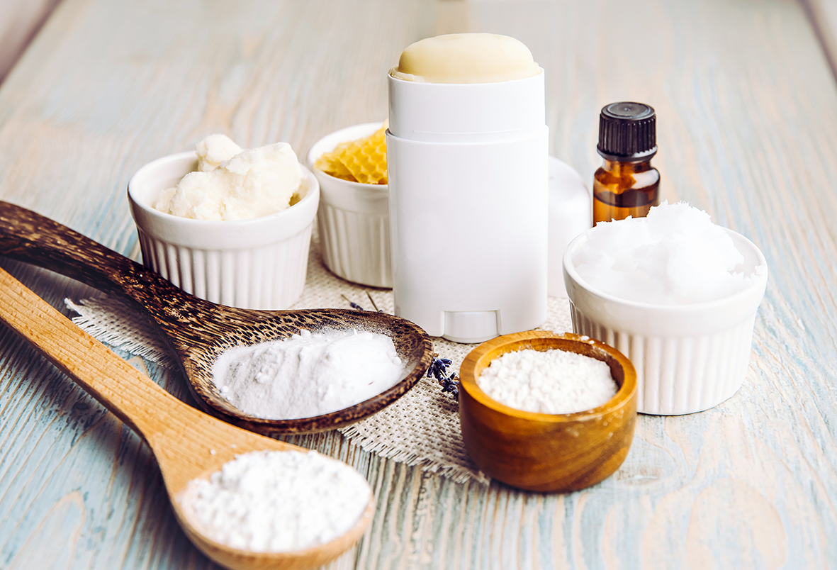 Solid deodorant on a light wooden table, accompanied by beeswax in a bowl and powdered raw materials on wooden spoons.